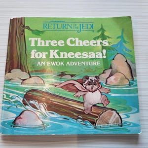 May include: A vintage Star Wars "Return of the Jedi" children's book. The cover features an Ewok character on a log in a river, with the title "Three Cheers for Kneesaa!" in white text against a green background.