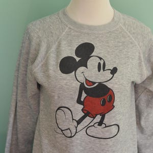 May include: A grey sweatshirt with a Mickey Mouse graphic. The cartoon character is in black, red, and white. The long-sleeved top is displayed on a mannequin.