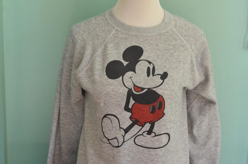 May include: A grey sweatshirt with a Mickey Mouse graphic. The cartoon character is in black, red, and white. The long-sleeved top is displayed on a mannequin.