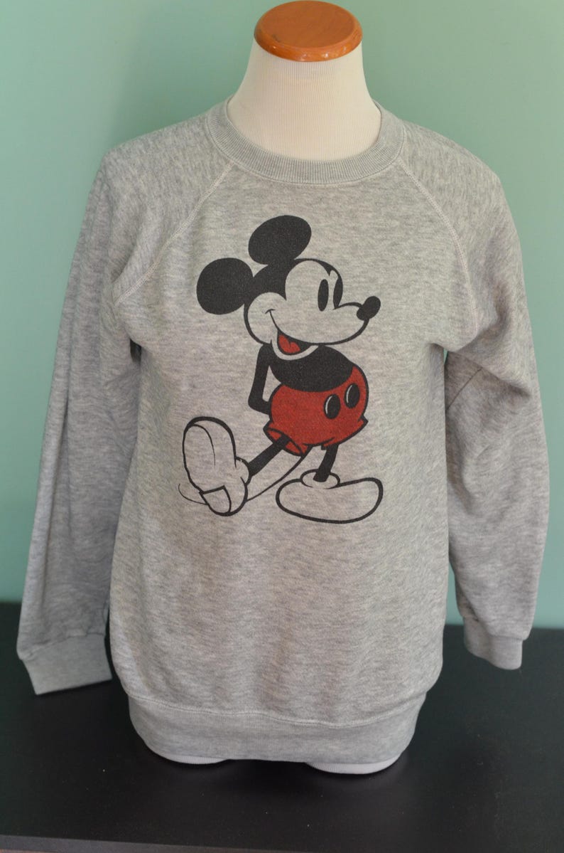 May include: A grey sweatshirt with a Mickey Mouse graphic. The cartoon character is in black, red, and white. The sweatshirt has long sleeves and a crew neck. A classic design.