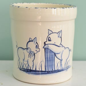 May include: A cylindrical ceramic crock with a cream-colored glaze and a blue hand-painted design featuring two pigs. One pig stands on the left, while the other leans against a post on the right. The rim has a distressed blue painted edge.