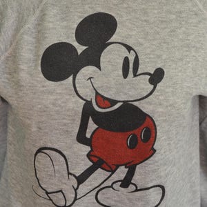May include: A grey sweatshirt with a large Mickey Mouse graphic. The cartoon character is in black, red, and white, smiling. The sweatshirt has a crew neck and raglan sleeves. A classic design.