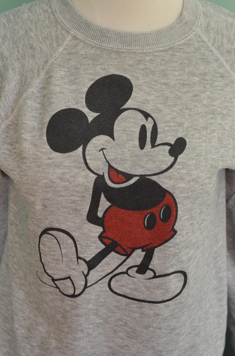 May include: A grey sweatshirt with a large Mickey Mouse graphic. The cartoon character is in black, red, and white, smiling. The sweatshirt has a crew neck and raglan sleeves. A classic design.