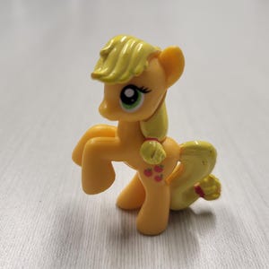 May include: A small yellow plastic pony figurine with a light yellow mane and tail. The pony has large green eyes, a yellow forelock, and a cutie mark on its flank featuring three small red apples. The figure is standing in a dynamic pose.