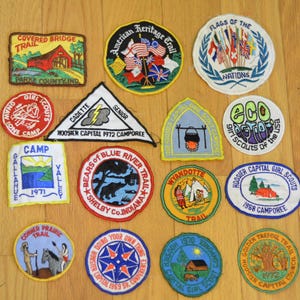 May include: A collection of 14 colorful embroidered patches featuring various designs, including a covered bridge, a girl scout camp, a blue river trail, and a golden tree. The patches are all circular or oval in shape and have a variety of colors and designs.