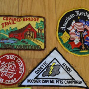 Peut inclure: Une collection de neuf patchs brod&eacute;s pr&eacute;sentant divers motifs. Les patchs sont color&eacute;s et pr&eacute;sentent des images d'un pont couvert, d'un camp de scouts f&eacute;minins, d'un &eacute;clair et de drapeaux am&eacute;ricains. Les patchs sont tous de forme rectangulaire ou circulaire et ont un fond blanc avec des contours noirs.