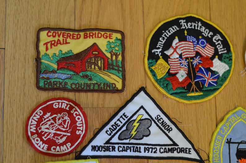 Peut inclure: Une collection de neuf patchs brod&eacute;s pr&eacute;sentant divers motifs. Les patchs sont color&eacute;s et pr&eacute;sentent des images d'un pont couvert, d'un camp de scouts f&eacute;minins, d'un &eacute;clair et de drapeaux am&eacute;ricains. Les patchs sont tous de forme rectangulaire ou circulaire et ont un fond blanc avec des contours noirs.