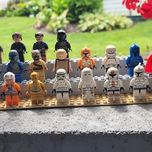 May include: A collection of twenty plastic minifigures arranged in three rows on a tan building block base. The figures feature various character designs including soldiers in white armor, a gold droid, an orange pilot, and figures in dark uniforms.