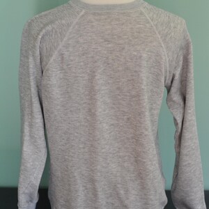 May include: A grey sweatshirt with long sleeves and a crew neck. The raglan sleeves are stitched with white thread. The sweatshirt is displayed on a white mannequin head and torso. The background is a light teal colour.