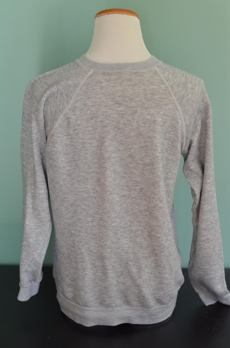 May include: A grey sweatshirt with long sleeves and a crew neck. The raglan sleeves are stitched with white thread. The sweatshirt is displayed on a white mannequin head and torso. The background is a light teal colour.