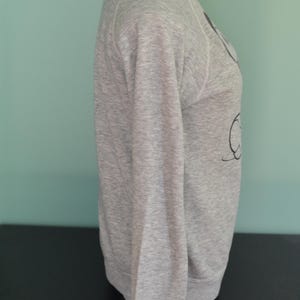 May include: A grey marl sweatshirt with long sleeves. The sweatshirt is displayed on a mannequin. The design on the front of the sweatshirt is a black graphic. The sweatshirt has a crew neck and ribbed cuffs.