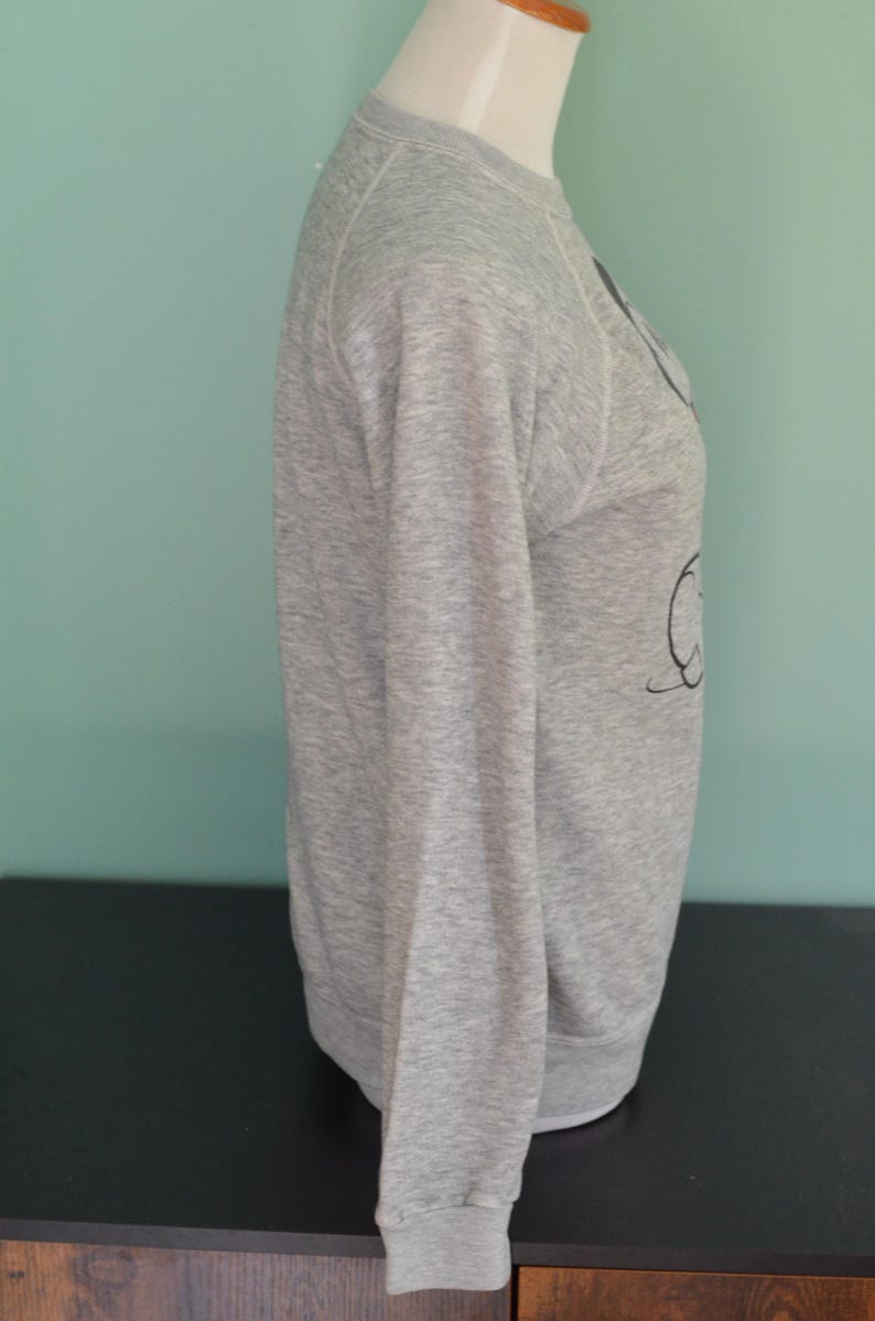 May include: A grey marl sweatshirt with long sleeves. The sweatshirt is displayed on a mannequin. The design on the front of the sweatshirt is a black graphic. The sweatshirt has a crew neck and ribbed cuffs.