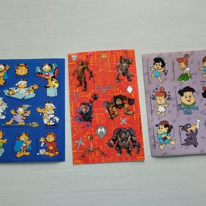 May include: Three sticker sheets featuring cartoon characters. One sheet is blue with Garfield in various costumes. Another is red with monster-like figures. The third is purple with characters from The Flintstones.