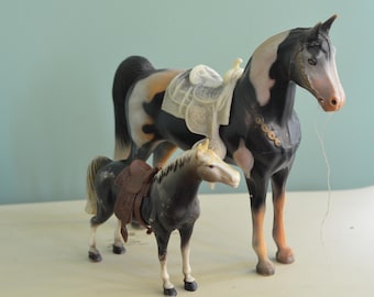 May include: Two toy horses, one black and white with a saddle and the other brown with a saddle. Both horses are standing on a white surface.