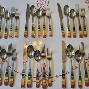 May include: A collection of stainless steel flatware with colorful striped handles. The set includes knives, forks, and spoons, arranged on a white surface. The handles feature bands of red, orange, yellow, green, blue, and purple.