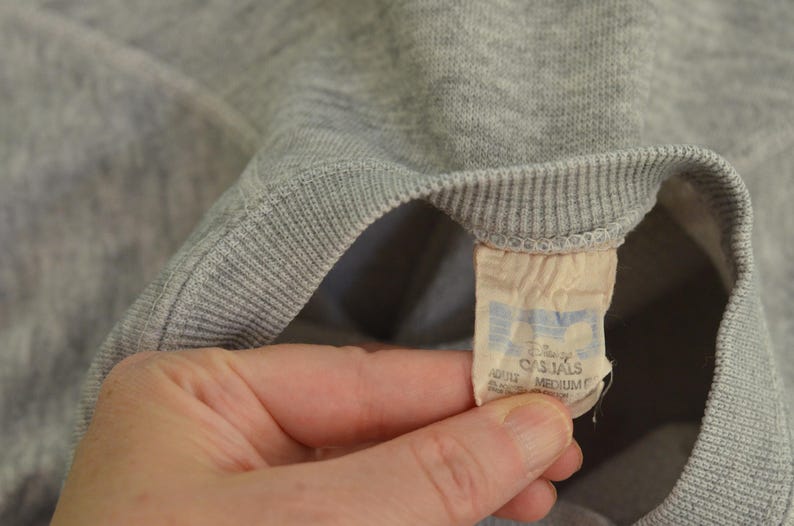 May include: Close-up of a grey sweatshirt with a ribbed collar and cuffs. A tag is visible, with the text "Disney CASUALS" and the size "MEDIUM". The sweatshirt is made of a soft, knit fabric.