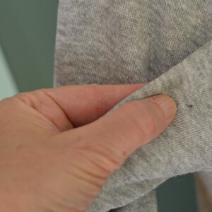 May include: Close-up of grey fabric, possibly a sleeve, held by a hand. The fabric has a slight texture and a small dark spot. The background is a muted green and white.