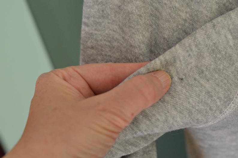 May include: Close-up of grey fabric, possibly a sleeve, held by a hand. The fabric has a slight texture and a small dark spot. The background is a muted green and white.