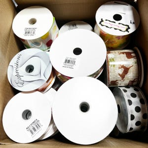 May include: A collection of ribbon spools in a cardboard box. The spools feature various designs, including polka dots, floral patterns, and solid white. Some spools have product information labels. The ribbons are suitable for crafts, gift wrapping, and decorative purposes.