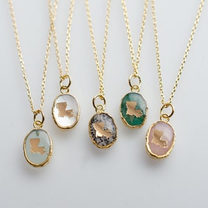 May include: Five gold necklaces with oval pendants featuring the state of Louisiana in gold on different colored backgrounds. The pendants are clear, green, gray, pink, and light blue.
