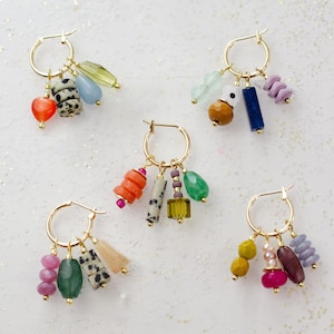 May include: Five pairs of gold hoop earrings, each adorned with colorful gemstone and bead charms. The charms include a variety of shapes and colors, such as red hearts, speckled beads, and green teardrops. The earrings are displayed on a white surface.