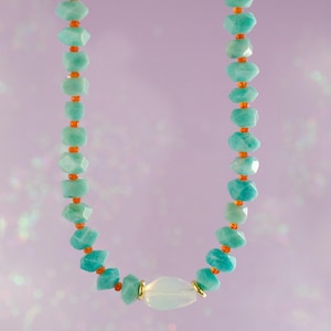 May include: A turquoise gemstone necklace with orange accent beads. The necklace features a large, oval, white gemstone in the center, with gold-colored metal accents. The necklace is displayed against a soft purple background.