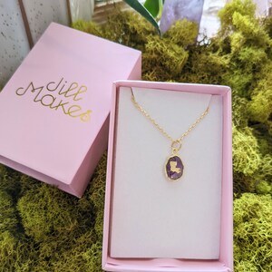 May include: A gold necklace with a purple pendant shaped like the state of Louisiana. The necklace is in a pink box with the words "Jill Makes" printed on the lid.