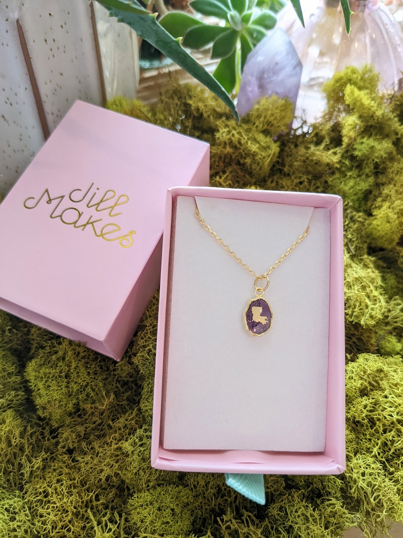May include: A gold necklace with a purple pendant shaped like the state of Louisiana. The necklace is in a pink box with the words "Jill Makes" printed on the lid.