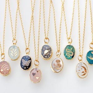 May include: Ten gold chain necklaces with oval-shaped pendants featuring various designs. The pendants are filled with resin and have gold accents. The designs include a leaf, a butterfly, a galaxy, a flower, a snake, a hand with a heart, a crescent moon, and a shooting star.