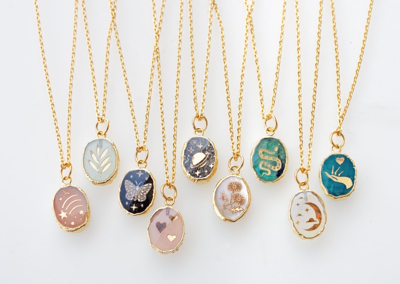 May include: Ten gold chain necklaces with oval-shaped pendants featuring various designs. The pendants are filled with resin and have gold accents. The designs include a leaf, a butterfly, a galaxy, a flower, a snake, a hand with a heart, a crescent moon, and a shooting star.