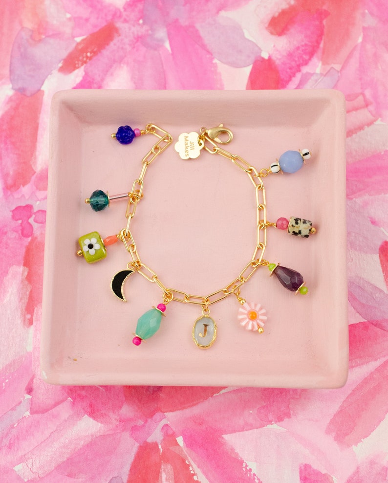 May include: A gold charm bracelet with a variety of colorful charms, including a flower, a crescent moon, a teardrop-shaped stone, and a charm with the letter "J". The bracelet is on a pink background.