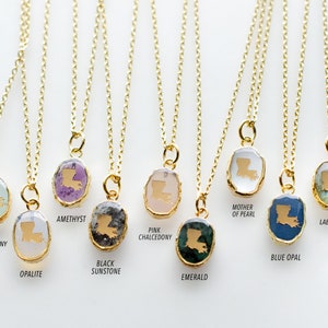 May include: Ten gold necklaces with pendants featuring the outline of the state of Louisiana. Each pendant is made of a different gemstone, including amethyst, aqua chalcedony, black sunstone, blue opal, emerald, labradorite, mother of pearl, opalite, and pink chalcedony.