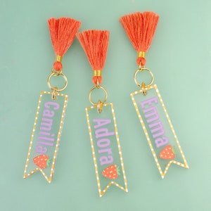 May include: Three clear acrylic keychains with gold rings and orange tassels. Each keychain has a white dotted border and a pink name, "Camilla", "Adora", and "Emma", with a small strawberry graphic.
