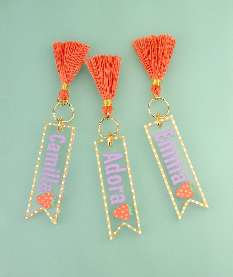 May include: Three clear acrylic keychains with gold rings and orange tassels. Each keychain has a white dotted border and a pink name, "Camilla", "Adora", and "Emma", with a small strawberry graphic.