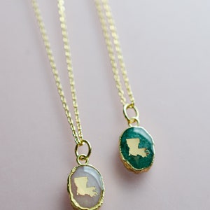 May include: Two gold necklaces with oval pendants featuring the state of Louisiana in gold foil. One pendant is set in a green stone, the other in a pink stone.