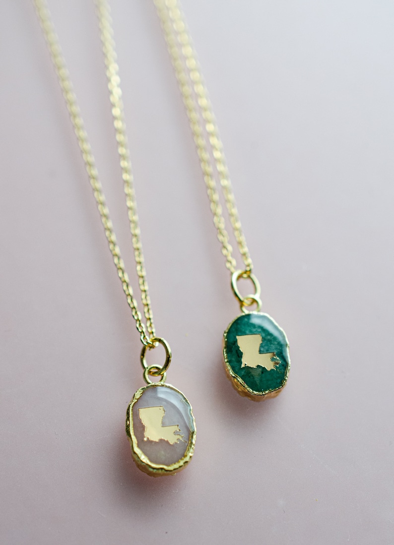 May include: Two gold necklaces with oval pendants featuring the state of Louisiana in gold foil. One pendant is set in a green stone, the other in a pink stone.