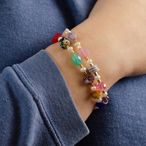May include: Two bracelets with colorful beads and pearls on a wrist. The bracelets are made with gold chain and have a variety of beads, including pink, green, blue, red, and white.