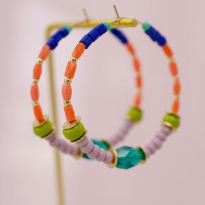 May include: A pair of hoop earrings with a multi-colored beaded design. The beads are pink, orange, green, blue, and purple. The earrings are hanging from a gold stand.