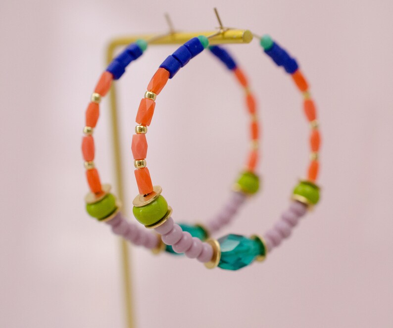 May include: A pair of hoop earrings with a multi-colored beaded design. The beads are pink, orange, green, blue, and purple. The earrings are hanging from a gold stand.