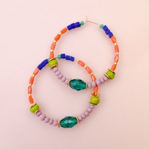 May include: A pair of hoop earrings with a multi-colored beaded design. The earrings are made with orange, green, blue, and purple beads, and feature two large, emerald green gemstone accents.