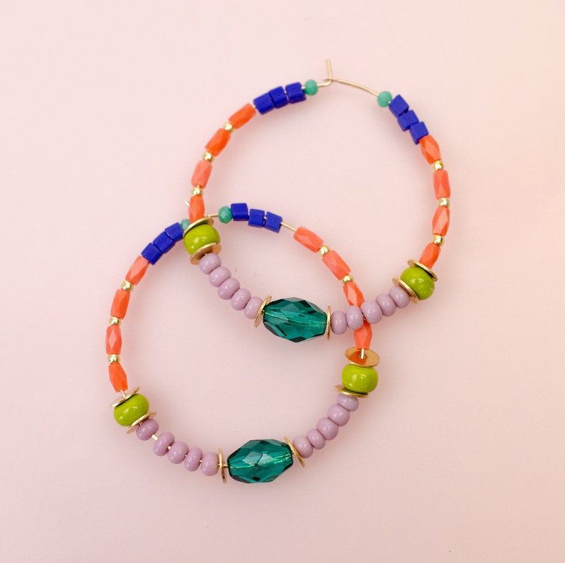 May include: A pair of hoop earrings with a multi-colored beaded design. The earrings are made with orange, green, blue, and purple beads, and feature two large, emerald green gemstone accents.
