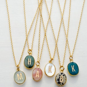 Op de afbeelding: Zes gouden kettingen met ovale hangers met verschillende letters en steenkleuren. De letters zijn H, A, K, B en K. De stenen zijn groen, roze, wit, zwart-wit en blauw.