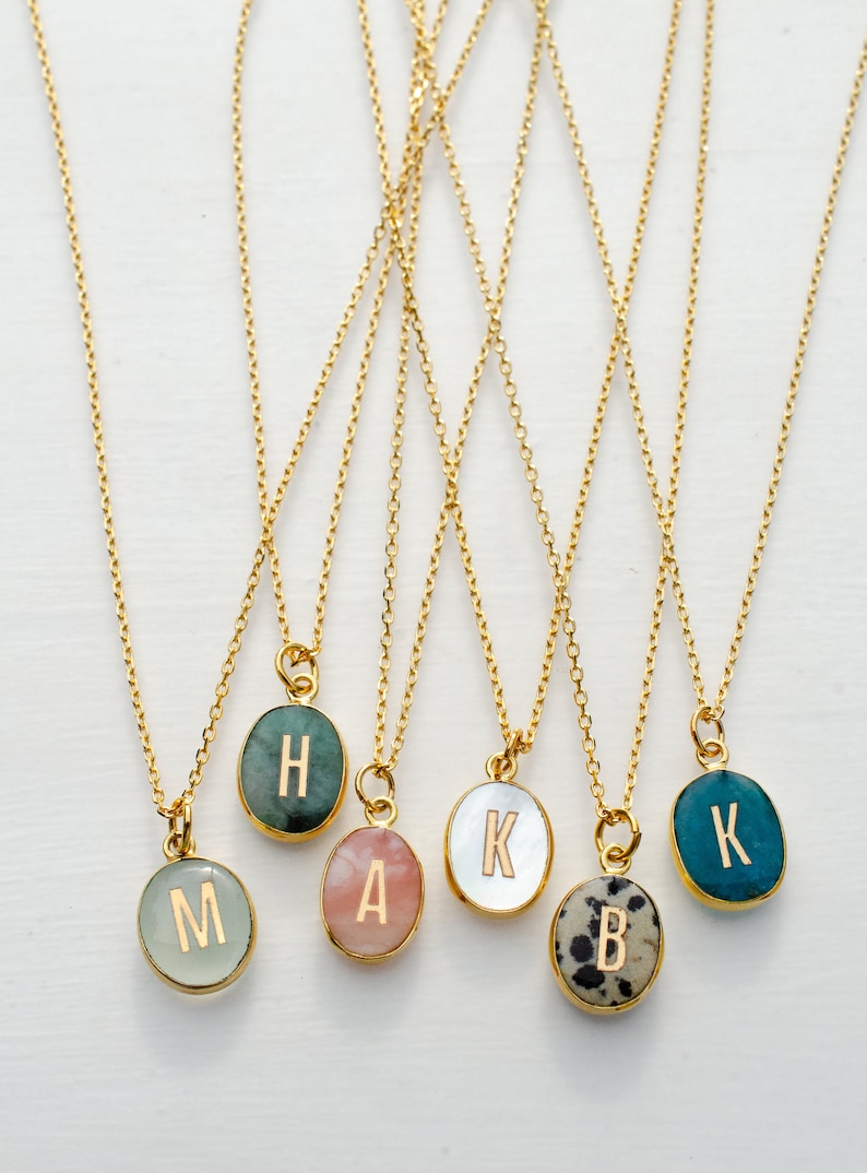 Op de afbeelding: Zes gouden kettingen met ovale hangers met verschillende letters en steenkleuren. De letters zijn H, A, K, B en K. De stenen zijn groen, roze, wit, zwart-wit en blauw.