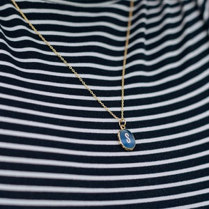 Op de afbeelding: Een gouden ketting met een blauwe ovale hanger met de letter 'S' op een zwart-wit gestreept shirt.