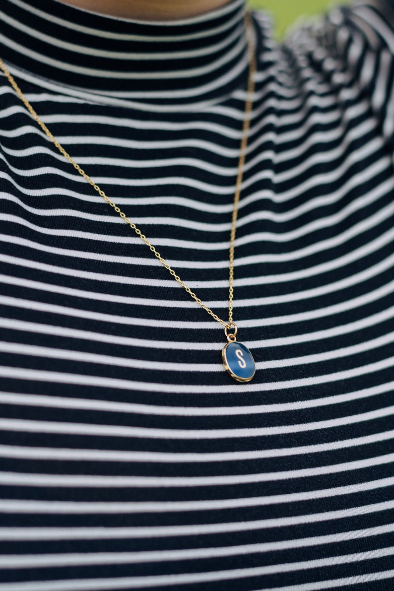 Op de afbeelding: Een gouden ketting met een blauwe ovale hanger met de letter 'S' op een zwart-wit gestreept shirt.