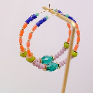 May include: A pair of hoop earrings with colorful beads. The earrings are made with orange, green, blue, and purple beads. The earrings are on a gold stand.