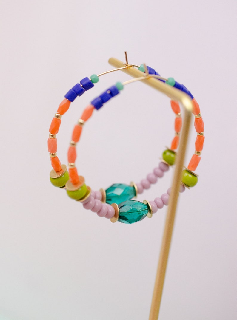 May include: A pair of hoop earrings with colorful beads. The earrings are made with orange, green, blue, and purple beads. The earrings are on a gold stand.