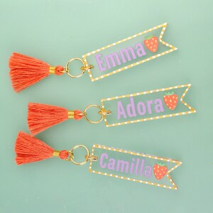 May include: Three keychains with orange tassels and clear acrylic tags with white polka dots and gold trim. The tags have the names "Emma", "Adora", and "Camilla" written in purple with a red strawberry graphic.
