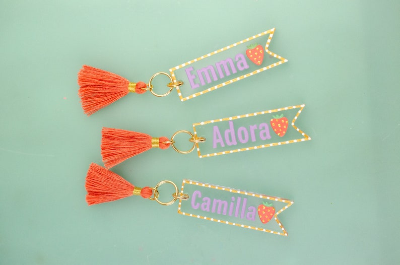 May include: Three keychains with orange tassels and clear acrylic tags with white polka dots and gold trim. The tags have the names "Emma", "Adora", and "Camilla" written in purple with a red strawberry graphic.