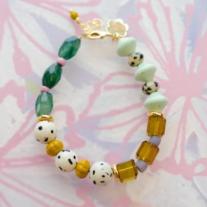May include: A colorful beaded bracelet featuring a mix of green, yellow, white, and light green beads. The bracelet includes oval green beads, speckled white beads, and gold-colored accents. The clasp is gold-toned.